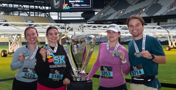 Los corredores lucen con orgullo sus medallas junto al trofeo del campeonato en el campo tras completar la carrera de 5 km «Por la salud mental infantil», lo que pone de relieve sus logros y el impacto en la comunidad.