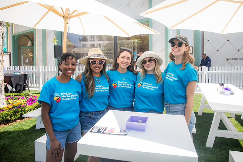 Un grupo de voluntarios sonrientes, vestidos con camisetas a juego de la Fundación para la Salud Mental Infantil, posan juntos bajo unas sombrillas blancas en un acto benéfico al aire libre.